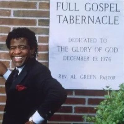 Al Green is standing with his hands on the wall in front of the board of the church.